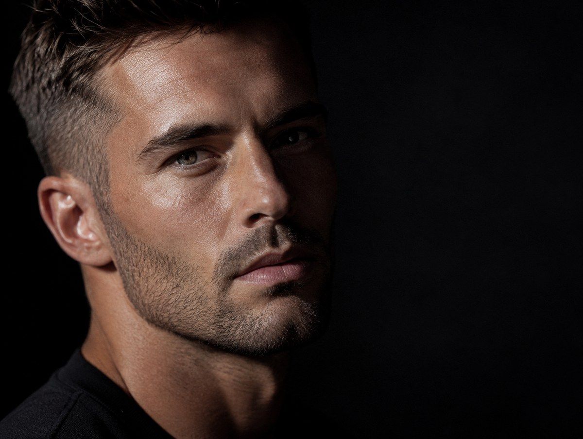 Man with short dark hair and stubble, side-lit against a dark background