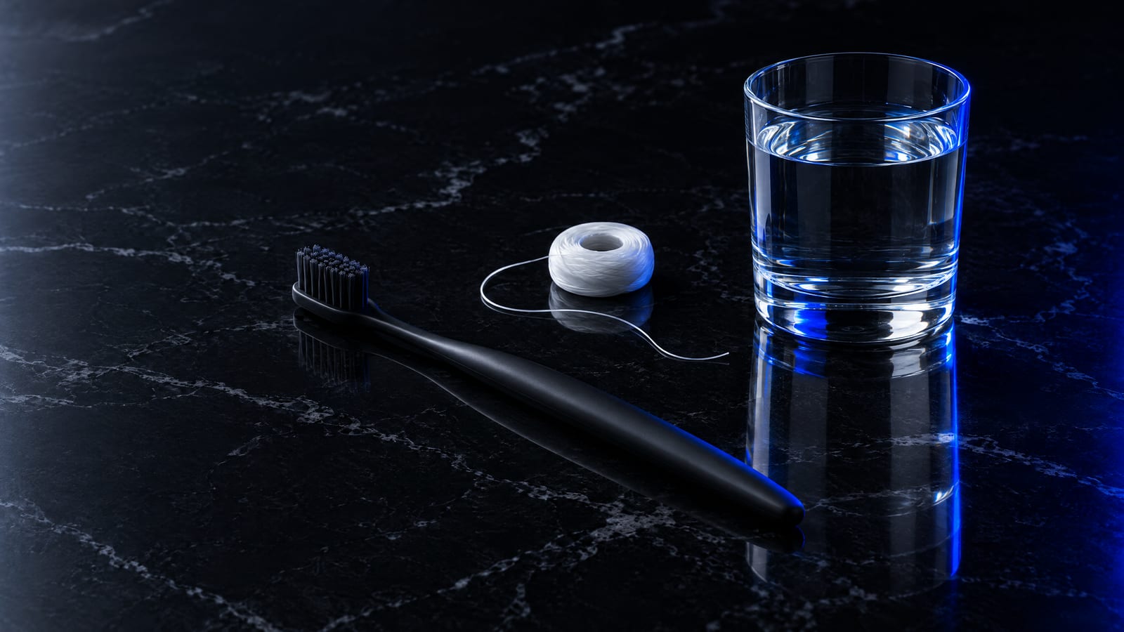 A black toothbrush, dental floss and a glass of water on black marble
